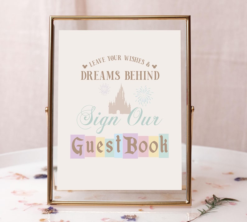 May include: A gold-framed sign with a white background and the text "Leave your wishes & dreams behind. Sign our guest book." The text is in a brown and blue font. The sign also features a silhouette of Cinderella's castle with fireworks.