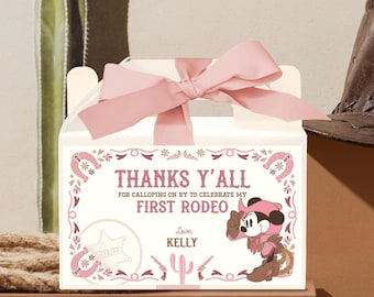 May include: A white gift box with a pink bow, featuring a rodeo theme. The box has text that reads "THANKS Y'ALL FOR CALLOPING ON BY TO CELEBRATE MY FIRST RODEO" with a cartoon character and sheriff badge.