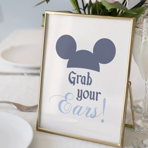 Puede incluir: Un cartel con marco dorado, fondo blanco y una silueta azul de Mickey Mouse. El cartel dice "Grab your ears!"