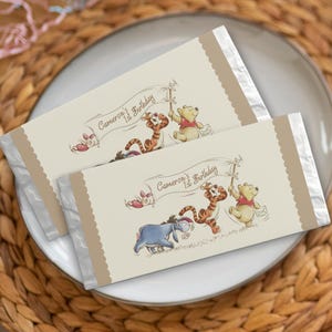 Peut inclure: Deux emballages de barres chocolatées sur le thème de Winnie l'Ourson. Les emballages sont beiges avec des extrémités en papier d'aluminium argenté et un bord festonné marron. Ils présentent des illustrations de Winnie, Porcinet, Tigrou et Bourriquet, avec le texte "Cameron's 1st Birthday".