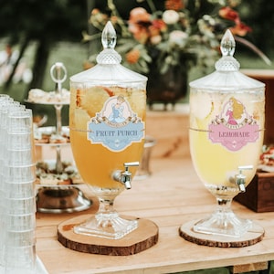Peut inclure: Deux distributeurs de boissons en verre remplis de punch aux fruits et de limonade. Chaque distributeur a un robinet et une étiquette avec une illustration de princesse et du texte. Le distributeur de punch aux fruits porte l'inscription "Cinderella's Fruit Punch".