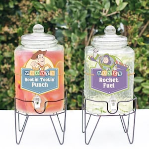 Op de afbeelding: Twee glazen drankdispensers met deksels, elk op een zwarte metalen standaard. Eén dispenser heeft een label met de tekst "Woody's Rootin' Tootin' Punch" en de andere "Buzz's Rocket Fuel". De dispensers zijn gevuld met kleurrijke dranken.