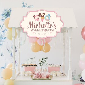 May include: A white dessert cart with a pink sign that says "Michelle's Sweet Treats Main Street" with a Mickey Mouse head and a cupcake, ice cream cone, and donut graphic. The cart is decorated with pink, white, and yellow balloons.
