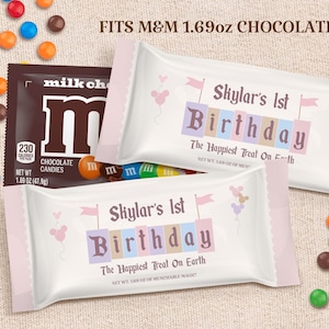 Könnte beinhalten: Ein weißer Schokoladenriegel-Wrapper mit rosa Besatz und einem rosa-blauen Geburtstagsdesign. Der Wrapper sagt "Skylar's 1st Birthday" und "The Happiest Treat On Earth". Außerdem steht "Fits M&M 1.69 oz Chocolate" darauf.