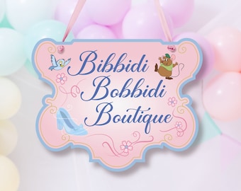 May include: A pink and blue sign with the text "Bibbidi Bobbidi Boutique" in blue cursive lettering. The sign features a blue bird, a glass slipper, and a mouse with a green hat.