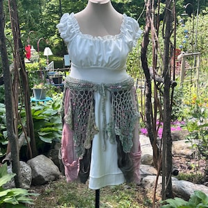 May include: A white dress with a square neckline and ruffled sleeves, paired with a crochet belt and a layered skirt in shades of pink and green. The dress is displayed on a mannequin in a garden setting, showcasing a bohemian or fairy-inspired aesthetic.
