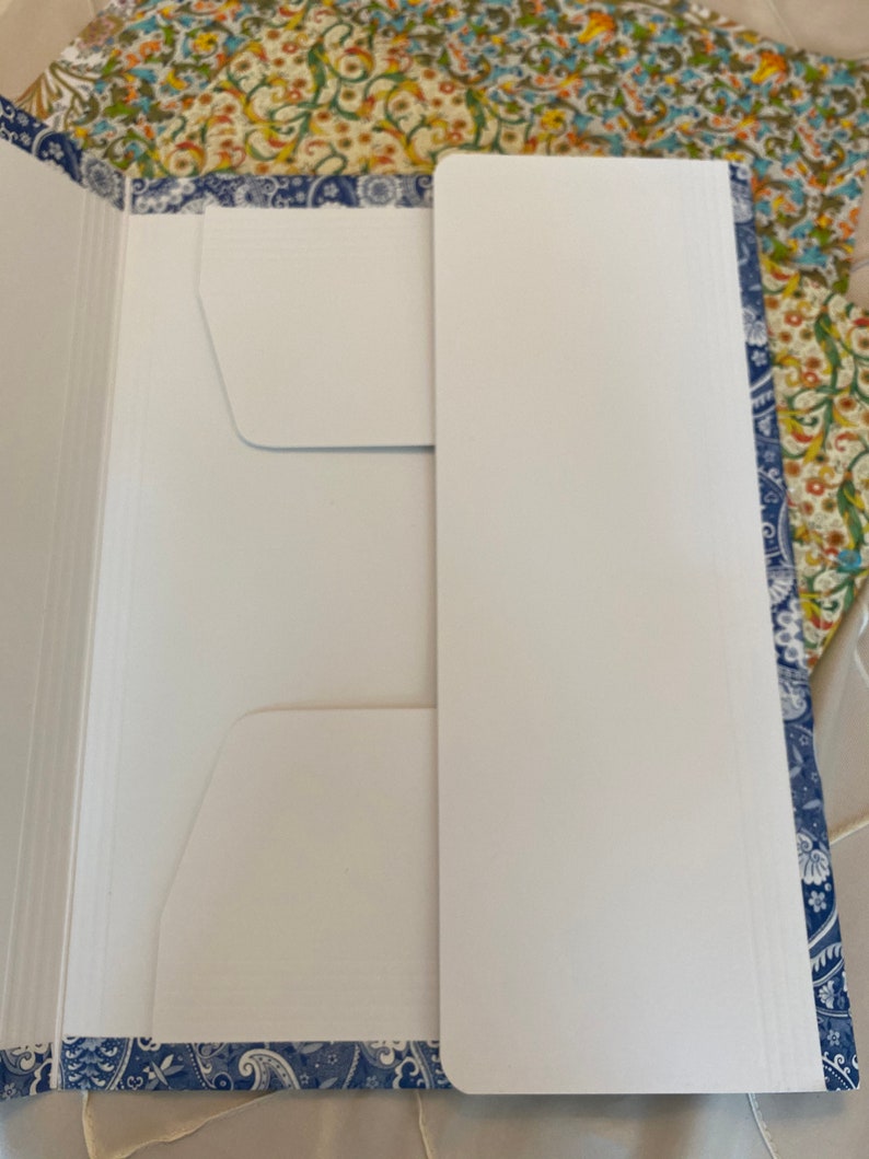 May include: An open white file folder with two pockets and a blue paisley border. The folder is on a floral patterned surface. The folder is designed to hold documents and papers.