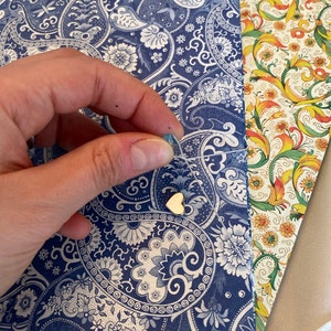 May include: A gold heart-shaped charm on a delicate string, set against a backdrop of patterned paper. The paper features a blue and white paisley design, with additional floral and solid-colored paper visible.