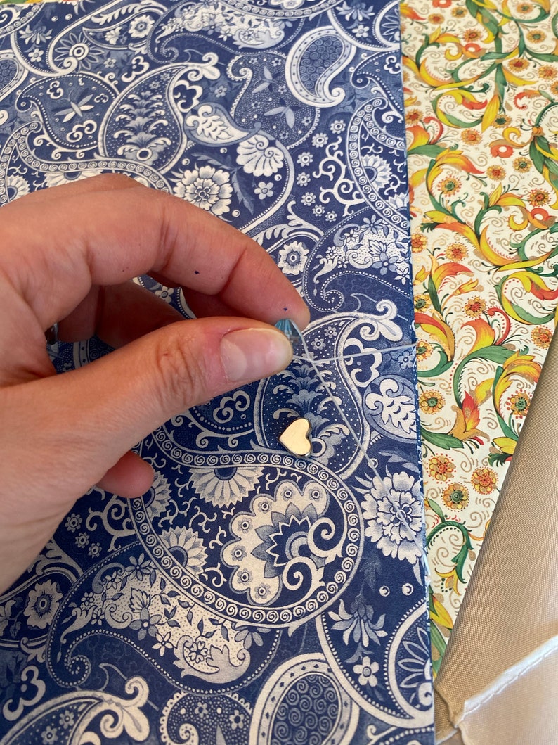 May include: A gold heart-shaped charm on a delicate string, set against a backdrop of patterned paper. The paper features a blue and white paisley design, with additional floral and solid-colored paper visible.