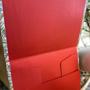 May include: Open view of a red paper folder with interior pockets. The folder has a decorative floral pattern on the edges and a gold-colored clasp. The interior is a solid, vibrant red.