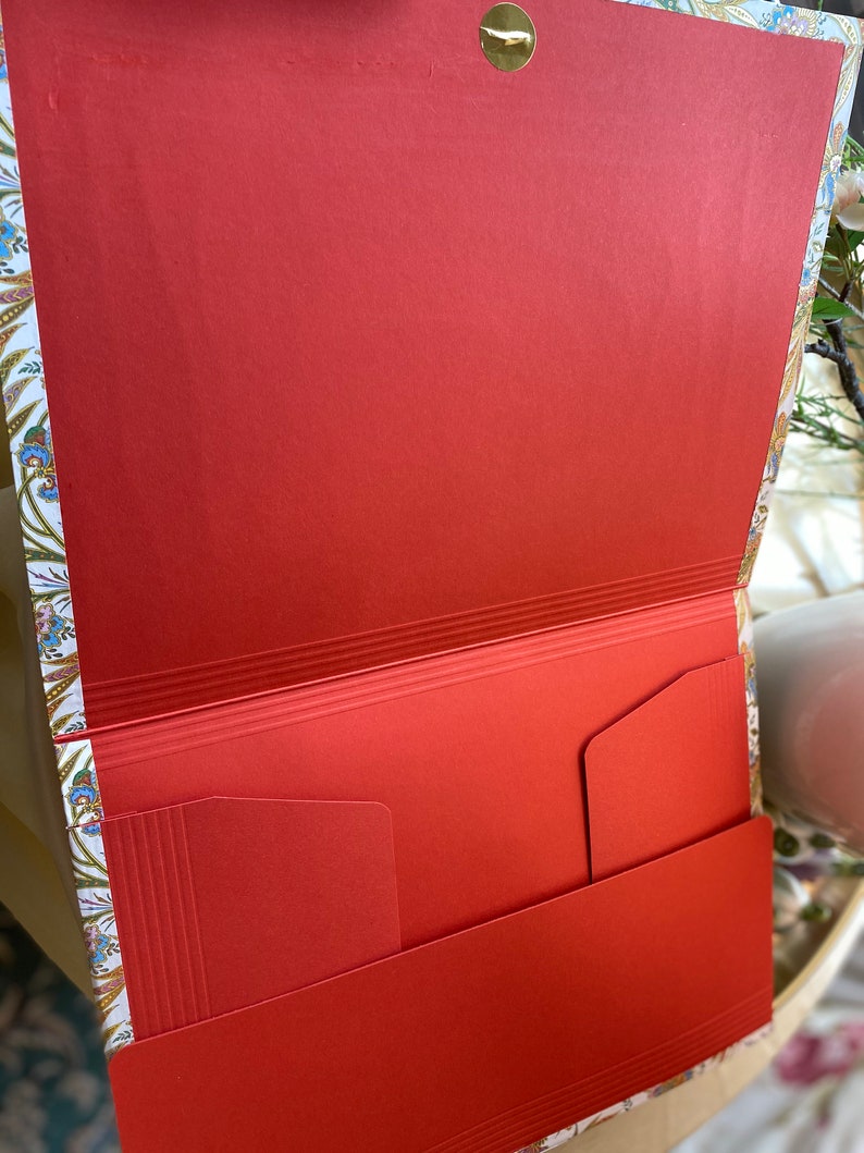 May include: Open view of a red paper folder with interior pockets. The folder has a decorative floral pattern on the edges and a gold-colored clasp. The interior is a solid, vibrant red.