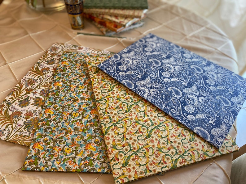 May include: Four decorative placemats with floral and paisley patterns in various colors. The placemats are arranged on a beige tablecloth. The patterns include shades of blue, green, yellow, and cream.