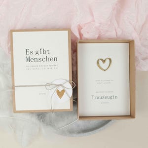 Könnte beinhalten: Eine Geschenkbox mit einer cremefarbenen Karte mit dem Text "Es gibt Menschen" und einem goldenen Herz. Die Box ist geöffnet und zeigt eine weitere Karte mit einem goldenen Herz und dem Wort "Trauzeugin". Die Box ist mit einer Schnur gebunden.