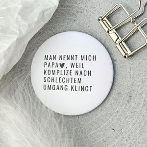 Könnte beinhalten: Ein weißer runder Button mit schwarzem Text: "MAN NENNT MICH PAPA, WEIL KOMPLIZE NACH SCHLECHTEM UMGANG KLINGT". Oben rechts befindet sich eine silberne Büroklammer. Oben links ist eine weiße Feder.