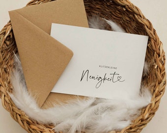 May include: A white card with the text "Klitzekleine" and "Heinigkeit" with a small heart symbol in a woven basket with white feathers.