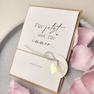 Könnte beinhalten: Eine weiße Grußkarte mit dem schwarzen Text "Für jetzt und für immer..." und "MARIE & PAUL". Die Karte ist mit einer dünnen braunen Kordel umwickelt, an der ein kleiner runder Anhänger mit einem goldenen Herz aus Folie befestigt ist.