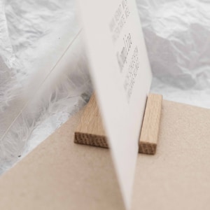 Puede incluir: Una tarjeta blanca con texto est&aacute; apoyada por dos bloques de madera. La tarjeta est&aacute; sobre una superficie marr&oacute;n.