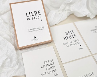 May include: A set of white milestone cards for pregnancy, with black text and a small box with a brown frame. The box reads "LIEBE IM BAUCH" and "30 MEILENSTEINKARTEN FÜR DEINE SCHWANGERSCHAFT". Several cards are visible, each with phrases like "SEIT HEUTE" and "DU BIST UNENDLICH GELIEBT".