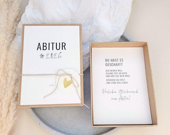 Puede incluir: Una caja de regalo de graduación con una tarjeta blanca que dice "ABITUR #2026" en texto negro. La tarjeta está atada con hilo y una pegatina de corazón dorado. La caja contiene una tarjeta con texto en alemán.
