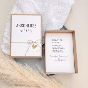 Könnte beinhalten: Zwei offene Geschenkboxen mit weißen Karten und braunem Karton. Die erste Box trägt das Wort "ABSCHLUSS" und die Nummer "#2026" auf der Karte. Die zweite Box trägt die Worte "DU HAST ES GESCHAFFT" auf der Karte.