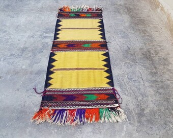 Puede incluir: Una alfombra larga de color amarillo con un patrón geométrico negro, rojo, verde y morado. La alfombra tiene flecos en ambos extremos.