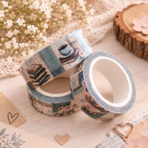 Könnte beinhalten: Zwei Rollen dekoratives Washi-Tape mit einem Vintage-inspirierten Design. Das Band zeigt Illustrationen von Bücherstapeln, einer Teetasse auf einer Untertasse und sanften Blumenmustern in gedämpften Blau-, Braun- und Cremetönen auf Papier.
