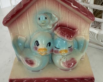 May include: A vintage ceramic birdhouse figurine featuring two blue birds nestled together, with a smaller bird above. The birdhouse has a red roof and base, with a cream-colored body. The birds have yellow beaks and red accents.