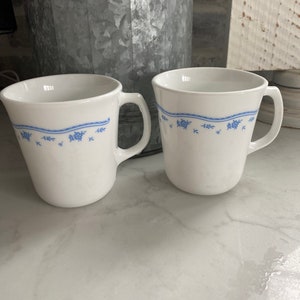May include: Two white ceramic mugs with blue floral designs and a blue trim around the top. The mugs have a handle on the right side.