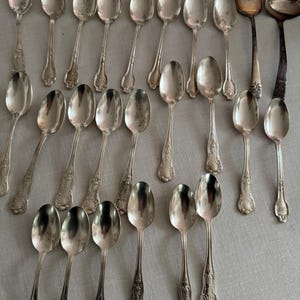May include: A collection of antique silver spoons, a fork, and a ladle arranged on a light gray surface. The spoons have ornate handle designs. The fork and ladle have a darker, tarnished appearance, contrasting with the spoons' brighter finish.