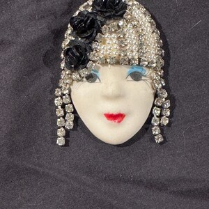 May include: A brooch featuring a porcelain face with painted blue eyeshadow, long eyelashes, and red lips. The figure wears a headpiece adorned with white faux pearls, clear rhinestones, and three black fabric flowers, with dangling rhinestone fringe.