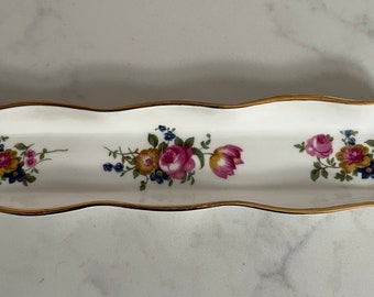 May include: A white porcelain trinket dish with a wavy edge and gold trim. The dish is decorated with a floral pattern in shades of pink, yellow, and blue.