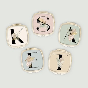 May include: Five compact mirrors with gold-coloured frames, each featuring a different letter: K, S, A, E, and T. The letters are black and decorated with botanical illustrations in neutral tones. The backgrounds are pastel colours.