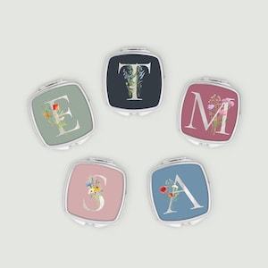 May include: Five square compact mirrors with floral initial designs. The letters are E, T, M, S, and A. Each mirror has a different background colour: sage green, navy blue, mauve, pink, and light blue. The letters are in a silver font.