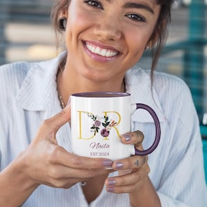 K&ouml;nnte beinhalten: Wei&szlig;er Keramikbecher mit lila Innenraum und Henkel. Der Becher zeigt goldfarbene Buchstaben, die "DR" mit floralen Akzenten buchstabieren, sowie den Namen "Naila" darunter und das Jahr "EST 2024".