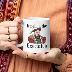 May include: White ceramic mug featuring a portrait of a historical figure with the text "It's all in the Execution". The portrait is in color, with the figure wearing a feathered hat and ornate clothing. The mug is held by a person.