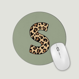 May include: A circular, sage green mouse mat featuring a leopard print letter "S". A white wireless mouse is positioned to the right. The mouse has a scroll wheel and two buttons. The mouse mat is approximately 20 cm in diameter.