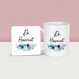May include: A white ceramic mug and matching square coaster. Both feature the text "Dr. Harriet" in black script, with a floral design in blue, pink, and teal. The mug has a handle on the right side.