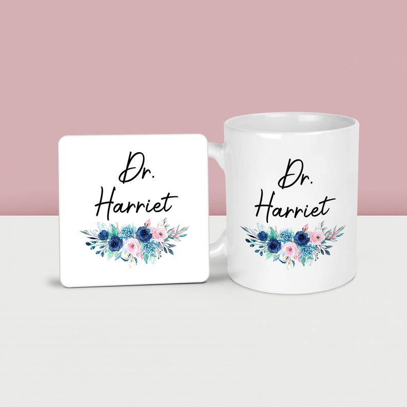 May include: A white ceramic mug and matching square coaster. Both feature the text "Dr. Harriet" in black script, with a floral design in blue, pink, and teal. The mug has a handle on the right side.