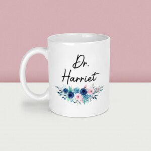 May include: White ceramic mug with the text "Dr. Harriet" in black script, adorned with a floral design in shades of blue and pink. The mug has a curved handle and is suitable for hot or cold beverages.