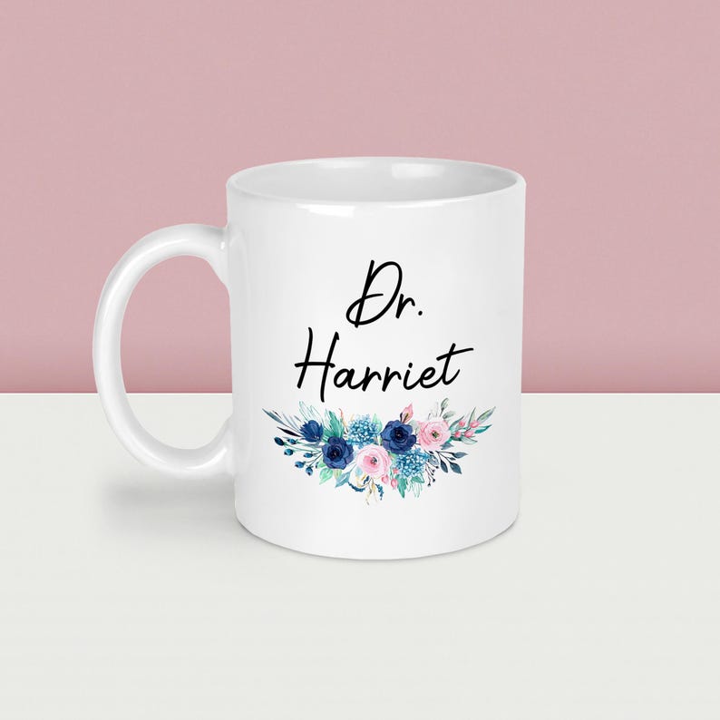 May include: White ceramic mug with the text "Dr. Harriet" in black script, adorned with a floral design in shades of blue and pink. The mug has a curved handle and is suitable for hot or cold beverages.