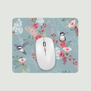 May include: A white wireless computer mouse sits on a floral mousepad. The mousepad has a light blue background with pink flowers, green leaves, and illustrations of hummingbirds and other birds.