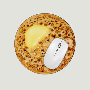May include: A circular mouse mat depicting a crumpet with melted butter, alongside a white wireless computer mouse. The mouse mat has a light beige background and a brown edge. The crumpet is a traditional British food.