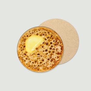 May include: A circular coaster depicting a crumpet topped with butter. The crumpet has a detailed, porous texture and a golden-brown colour. A second, plain, beige coaster is also visible.