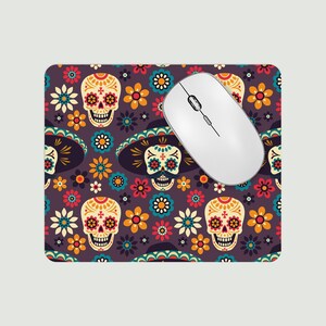 May include: A rectangular mouse mat with a vibrant Day of the Dead design. The pattern includes sugar skulls, sombreros, and flowers in orange, yellow, blue, and red on a dark purple background. A white wireless mouse is on the mat.
