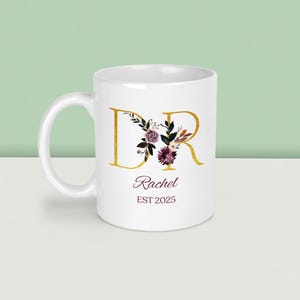 K&ouml;nnte beinhalten: Wei&szlig;e Keramiktasse mit goldenen Buchstaben "DR" und floralen Akzenten in Lila, Burgunderrot und Gr&uuml;n. Der Name "Rachel" und das Jahr "EST 2025" sind darunter aufgedruckt. Die Tasse hat einen gebogenen Henkel.