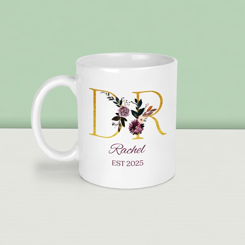K&ouml;nnte beinhalten: Wei&szlig;e Keramiktasse mit goldenen Buchstaben "DR" und floralen Akzenten in Lila, Burgunderrot und Gr&uuml;n. Der Name "Rachel" und das Jahr "EST 2025" sind darunter aufgedruckt. Die Tasse hat einen gebogenen Henkel.