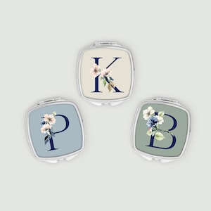 May include: Three square compact mirrors with floral initial designs. Each mirror features a different letter: K, P, and B, in navy blue, surrounded by delicate floral arrangements in shades of white, pink, and blue. The backgrounds are light beige, blue, and green.