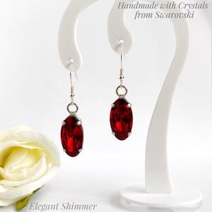May include: A pair of silver-toned dangle earrings with oval, deep red crystals. The earrings are displayed on a white stand, with the text "Handmade with Crystals from Swarovski" and "Elegant Shimmer".