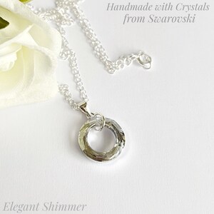 May include: A silver necklace with a circular pendant made with crystals from Swarovski. The pendant is a clear, faceted ring. The text "Handmade with Crystals from Swarovski" and "Elegant Shimmer" are visible.
