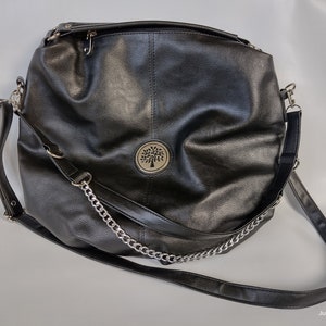 Könnte beinhalten: Eine schwarze Ledertasche mit einem silbernen Kettenriemen und einem runden Logo mit einem Baummotiv.